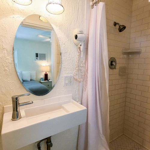 A bathroom with a sink, mirror, hairdryer, and shower with a curtain; a reflection of a bedroom is visible in the mirror.