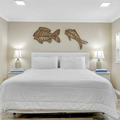 A bedroom with a large bed, two lamps on nightstands, and wall art of fish above the bed in a neat, coastal style.