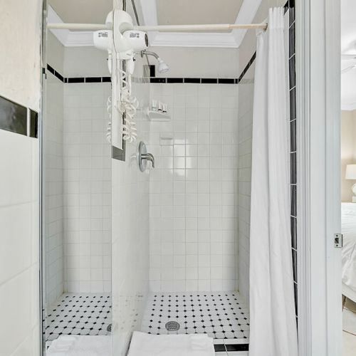 The image shows a white-tiled shower with a curtain, next to a bedroom with a neatly made bed and ceiling fan.