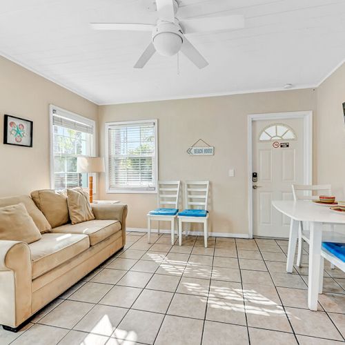 A cozy living room with a beige sofa, dining table, wall art, and a TV. Light walls, tiled floor, and abundant natural light complete the space.