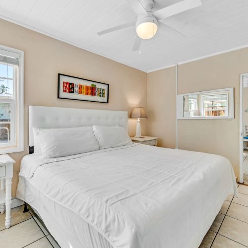 A cozy bedroom with a white bed, bedside tables, art on the wall, a window, a mirror, and a door leading to another room.