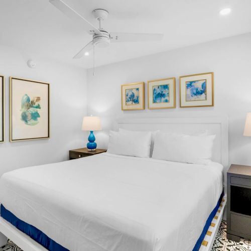 A modern bedroom with a bed, blue lamps, artwork on the walls, and a ceiling fan, featuring white and blue decor.