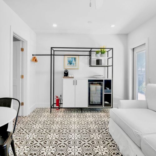 A modern room with patterned floor tiles, white sofa, dining table, mini fridge, and wall art of a beach scene. Brightly lit space.