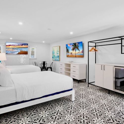 The image shows a bright room with two beds, patterned floor, wall art, a shelving unit, and a small fridge.