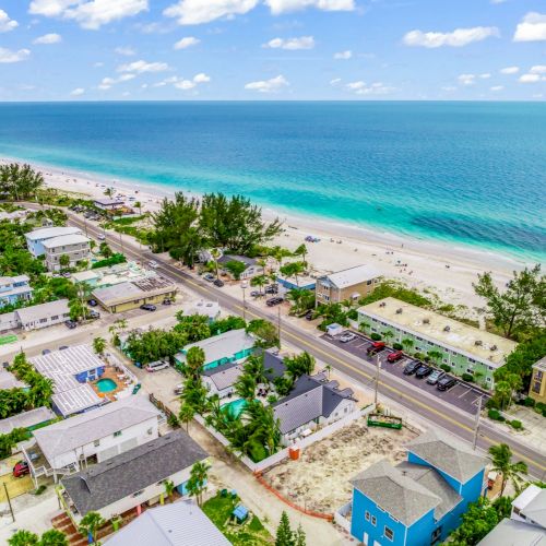 Coastal townscape with houses, a road, and parked cars near a sandy beach and azure ocean under a partly cloudy sky.