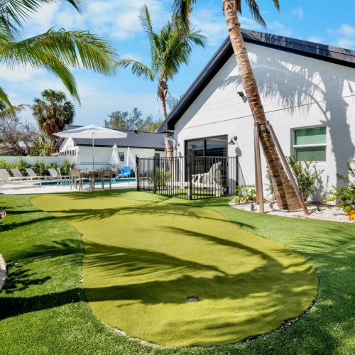 A backyard features a putting green, palm trees, and a swimming pool next to a modern house under a clear blue sky.