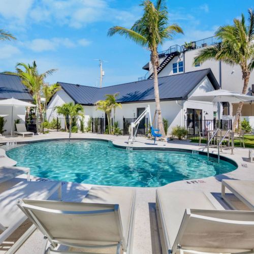 The image shows a resort pool with lounge chairs, umbrellas, and palm trees under a clear blue sky, creating a relaxing tropical setting.