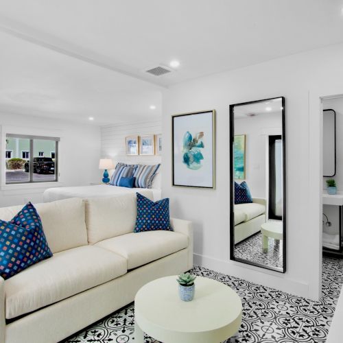 A modern living room with a white couch, patterned pillows, wall art, a mirror, small round table, and a glimpse of a bathroom beyond a door.