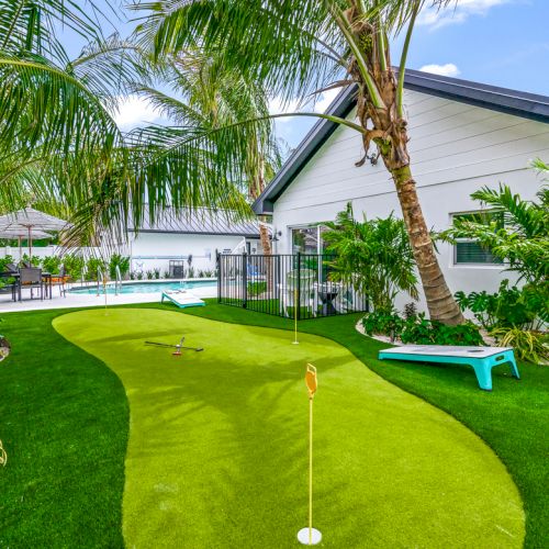 A backyard features a mini-golf course with lush greenery and palm trees, and a poolside area next to a modern house.