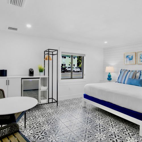 The image shows a bright, modern hotel room with a bed, a table with chairs, a TV, and decorative elements on the walls.