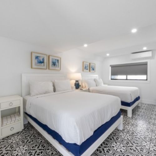 A bright bedroom features two beds, blue accents, patterned flooring, nightstand, lamps, three paintings, and a window with a blind.