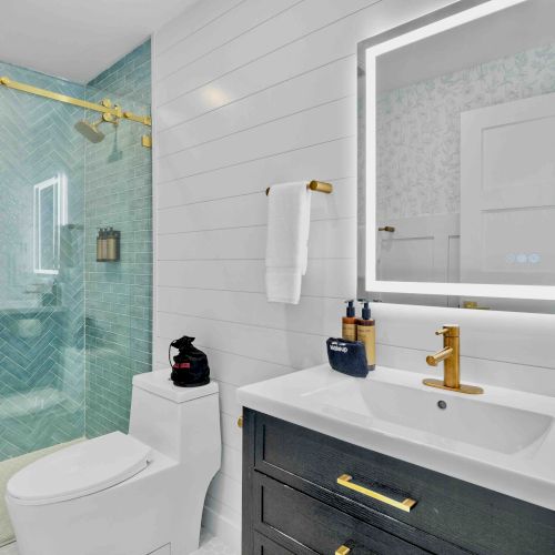 A modern bathroom with a glass shower enclosure, a white toilet, a dark vanity with gold hardware, a backlit mirror, and white tiled walls.