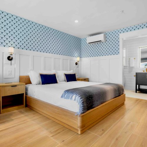 A bright hotel room with a wooden platform bed, blue and white bedding, a blue armchair, nightstands, patterned blue wallpaper, and a small sink area in a doorway.