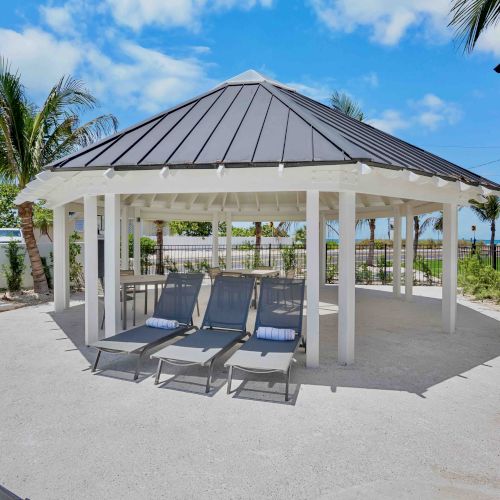 A modern outdoor pavilion with glass walls, a shaded seating area, tiled walkway, and bright blue sky behind.