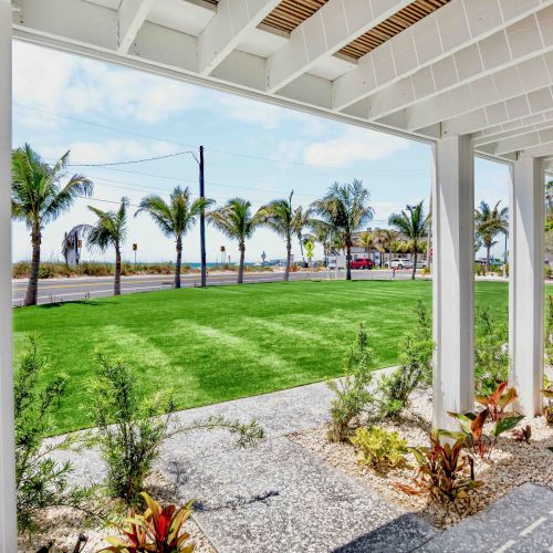 A sunny porch overlooks a bright green lawn and palm trees, with a table and chairs, and a calm open view beyond the railing.
