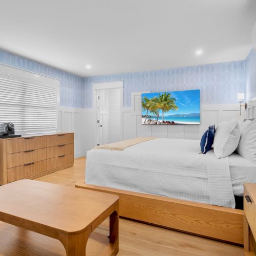 A bright hotel room with a wooden bed frame, white bedding, light blue walls, a dresser, desk, and a small table, plus a tropical TV image on the wall, ending with a period.