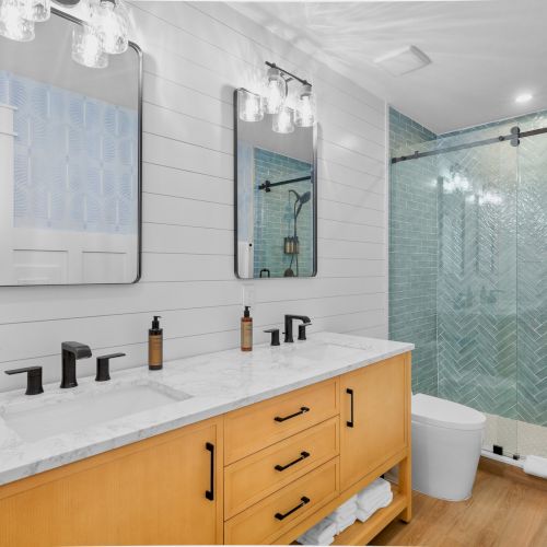 A modern bathroom with a double vanity, light wood cabinets, white countertops, black faucets, two mirrors, and a glass shower with a sliding door in a herringbone-tiled blue-green shower wall.
