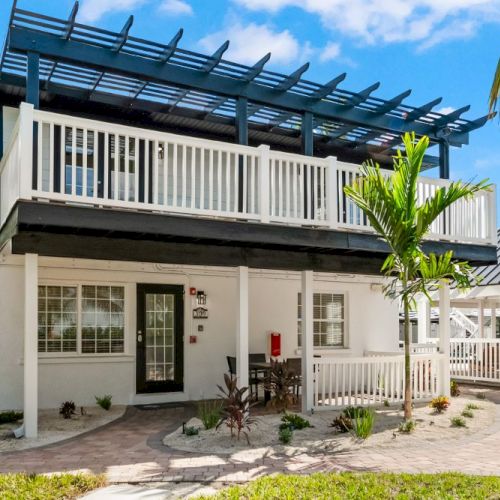 A tropical-style house with white railings, surrounded by palm trees and a gazebo on a sunny day.
