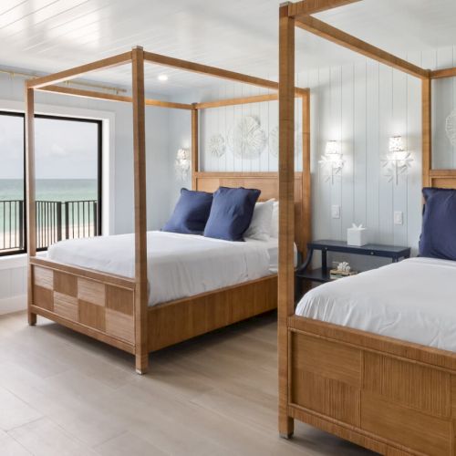 A bright bedroom with two canopy beds, ocean view from a large window, modern decor, light wooden floor, and stylish furnishings.