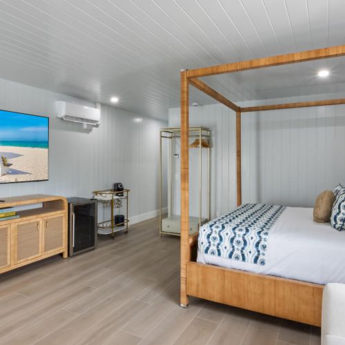 The image shows a bright bedroom with a canopy bed, flat-screen TV, and a wooden console, decorated in a coastal style.