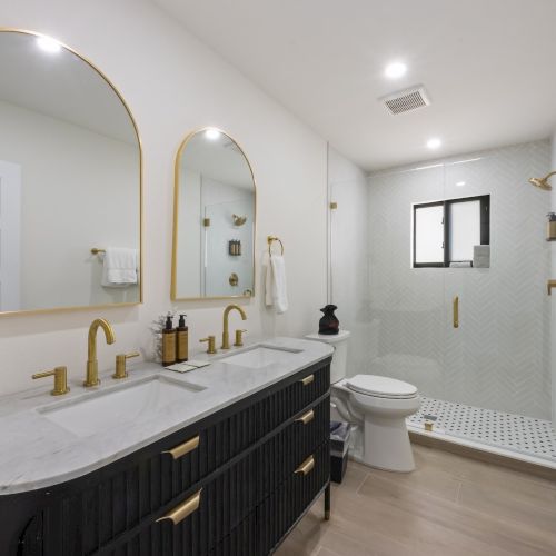A modern bathroom with dual sinks, gold fixtures, a glass shower, and a towel rack on a white wall, illuminated by recessed lighting.