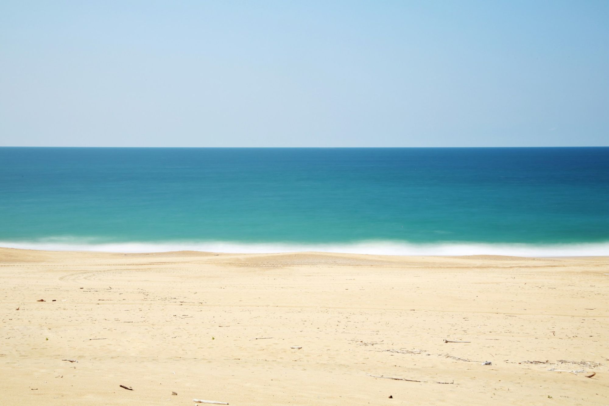 A tranquil beach scene with pale sand and calm turquoise-blue ocean under a clear blue sky, creating a serene and peaceful atmosphere.