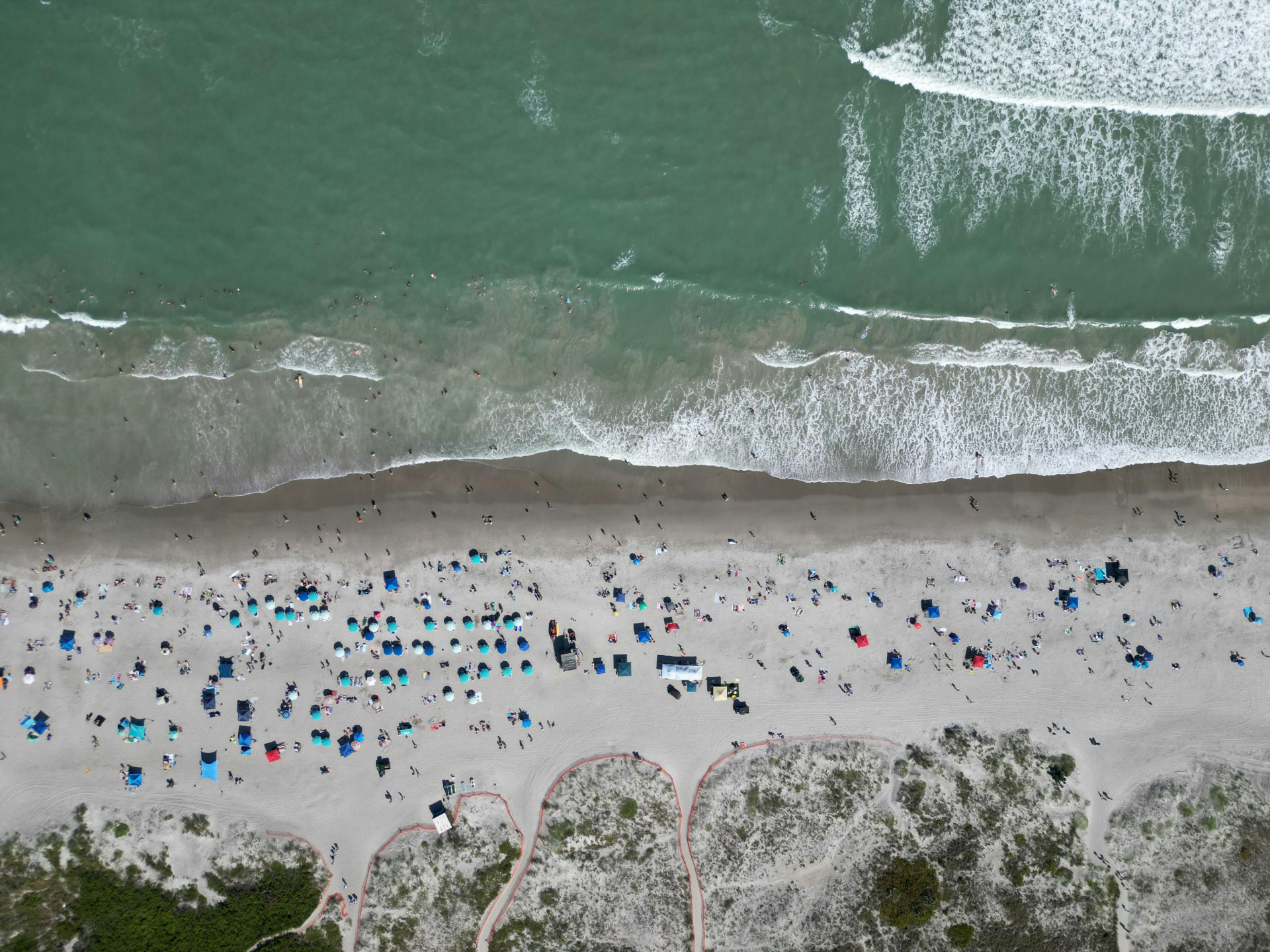 An aerial view of a beach with waves, many people, and colorful umbrellas scattered across the sand.