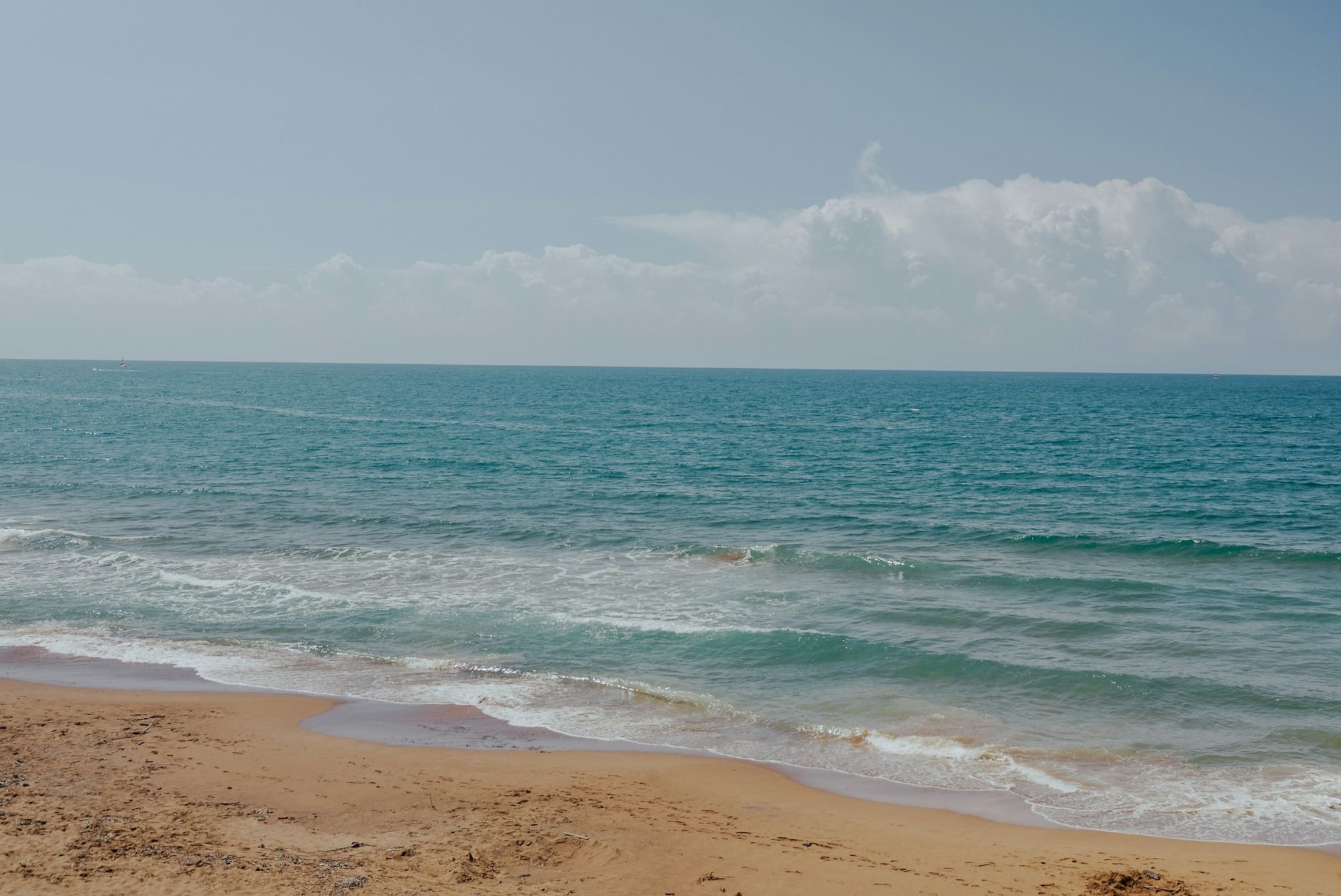 A sandy beach with gentle waves and a clear blue sky above the sea horizon creates a tranquil scene.