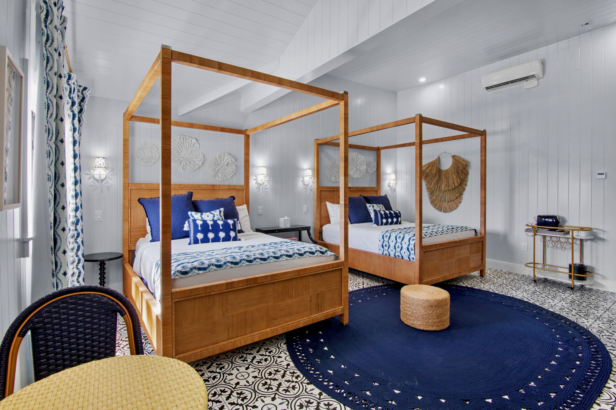 Two canopy beds with blue bedding, a round rug, wicker stools, patterned floor, and a cozy, coastal-themed decor in a bright room.