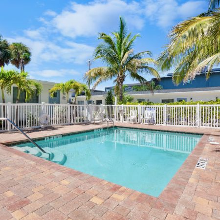 A small outdoor swimming pool with a white fence, surrounded by palm trees and a building under a blue sky.