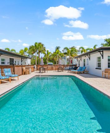 A swimming pool with lounge chairs and striped umbrellas, surrounded by white buildings, set against a backdrop of palm trees and blue sky.