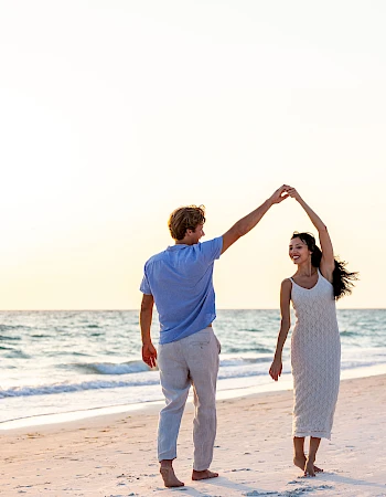 A couple is dancing joyfully on a sandy beach by the ocean, enjoying a beautiful sunset in the background.