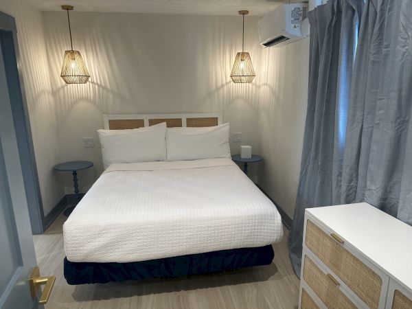 A cozy bedroom with a neatly made bed, two side tables, hanging lamps, a dresser, and curtains, all in a neutral color palette.