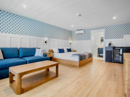 A bright, open hotel suite with a blue and white color scheme, featuring a blue sectional sofa, a wooden coffee table, a king bed, wallpapered walls, tiled accents, and a small kitchenette area. Always ending the sentence.