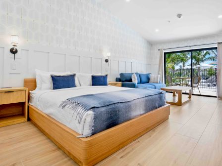 A bright hotel room with two large beds, blue accents, wooden furniture, and a balcony overlooking palm trees and a sunny outdoor view.