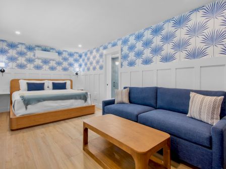 A bright bedroom with a wooden bed, blue upholstered sofa, a matching blue-and-white patterned wall, and a wooden coffee table, light wood floors, and white wainscoting, with bedside tables and lamps.