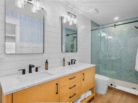 A modern bathroom with a double sink vanities, light wood cabinet, white countertop, two mirrors, and a glass shower with a blue chevron tile pattern. The room is bright and clean.