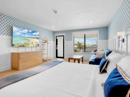 A bright hotel-like bedroom with two queen beds, a blue and white color scheme, wall sconces, a wall-mounted TV, a sofa, coffee table, dresser, and large windows showing a sunny ocean view, ending now.