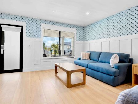 A bright living room with a blue patterned wallpaper, white wainscoting, a blue sofa, a wooden coffee table, and a window letting in light.