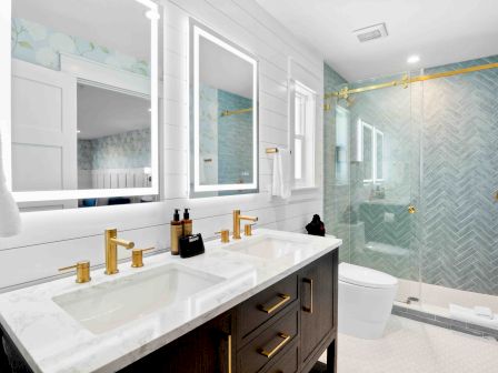 A modern double-sink bathroom with a white marble countertop, gold fixtures, a dark vanity, triple mirrors, and a glass-enclosed shower.