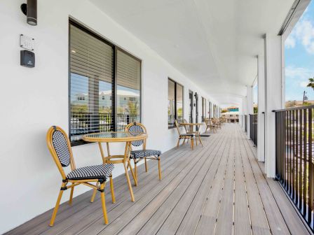 A sunny outdoor hotel balcony with small round tables and wicker chairs along a long wooden deck, white walls, windows, and a railing&mdash;calm resort vibe.