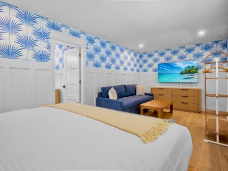 A bright hotel-style room with a white bed, blue palm-pattern wallpaper, a blue sofa, a wooden dresser with a TV, and a wooden rack, all on a warm wooden floor.