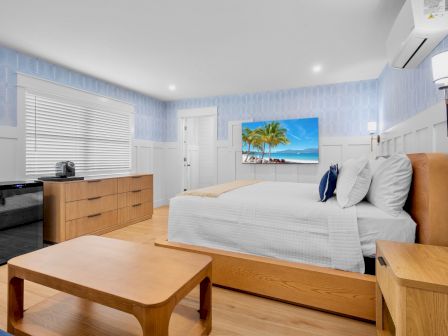 A bright hotel room with a large bed, wooden furniture, a desk with a kettle, a wall-mounted TV showing a tropical beach, and blue wallpaper, ending with a period.