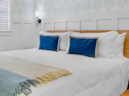 A neatly made bedroom with a white bed, wooden headboard, two blue pillows, a pale yellow/blue throw, and soft wall lighting by the window, immaculate and bright.