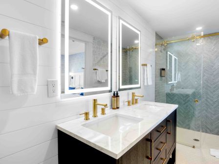 A modern bathroom with a double vanity, gold fixtures, white countertops, mirrored cabinets, towel rack, and a glass-enclosed tiled shower, bright and clean.