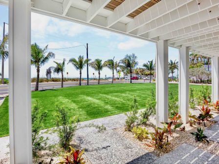 A covered porch overlooks a well-kept lawn with palm trees, gravel borders, and tropical plants, walking path, and a sunny coastal scene in the background.