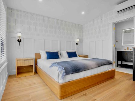 A bright, modern hotel-style bedroom with a wooden platform bed, white paneled walls, blue accents, and a small desk area by the door.