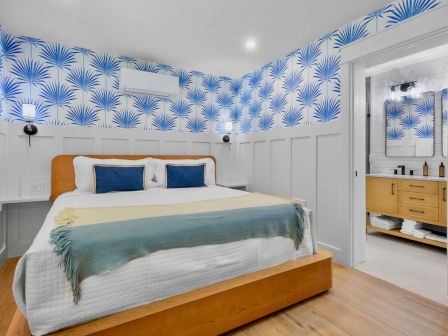 A cozy hotel-style bedroom with a wooden bed, blue palm-patterned wallpaper, white paneling, light wood floors, and a bright en-suite bath.