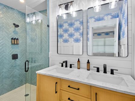 A modern bathroom with a glass shower, blue tiled walls, a white marble countertop, black faucets, and wooden vanity drawers beneath two mirrors with blue palm-patterned wallpaper.