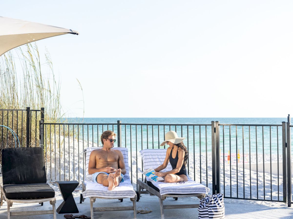 A man and a woman relax on lounge chairs by a pool with an ocean view, under sunny skies.